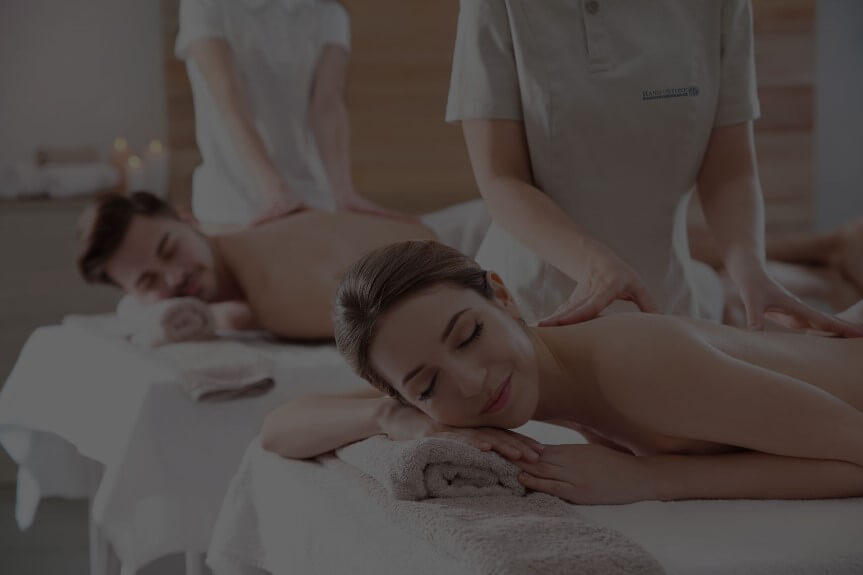 Two people lying with eyes closed, heads resting on small pillows as they receive massages.